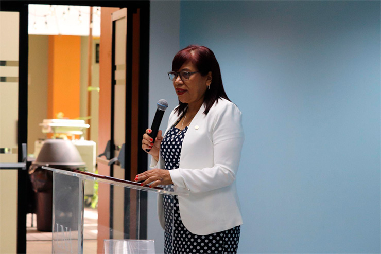 La imagen muestra a la vicerrectora de extensión, Yolanda Pérez tras un pulpito, con un micrófono en la mano, participando del evento.